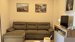 Sala de estar con un sofá modular, una mesa de centro con pasteles y dos tazas de café. Hay cuadros en la pared y un televisor sobre un mueble de madera.