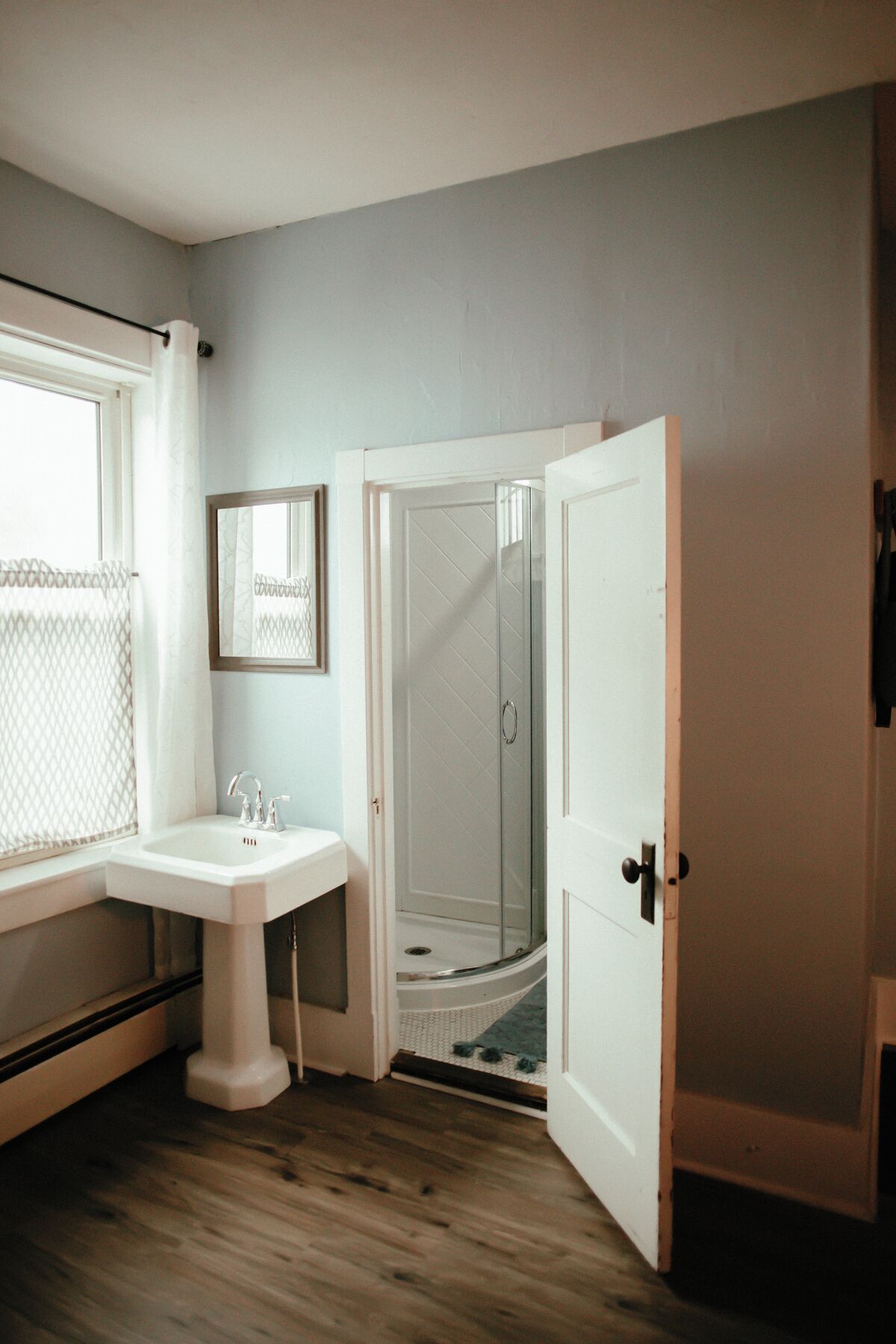 A bathroom with a white pedestal sink, a glass shower enclosure, and a door leading to another room.