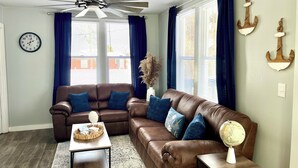 A living room with a brown leather sofa, a coffee table, and a ceiling fan. There are two windows with blue curtains and a wall clock. - Charming 2-bedroom house in serene Coloma with AC, WiFi (Coloma)