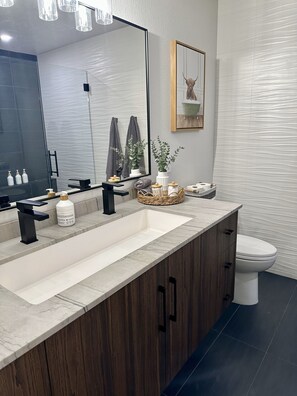 A modern bathroom with a large mirror, a sink with a wooden cabinet, a toilet, and a small plant on the counter. - Charming 3-bedroom House in Queen Creek w/ Community Pool (Queen Creek)