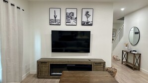 A living room with a large flat-screen TV mounted on the wall, wooden entertainment unit, and a wooden coffee table. - Charming 3-bedroom House in Queen Creek w/ Community Pool (Queen Creek)