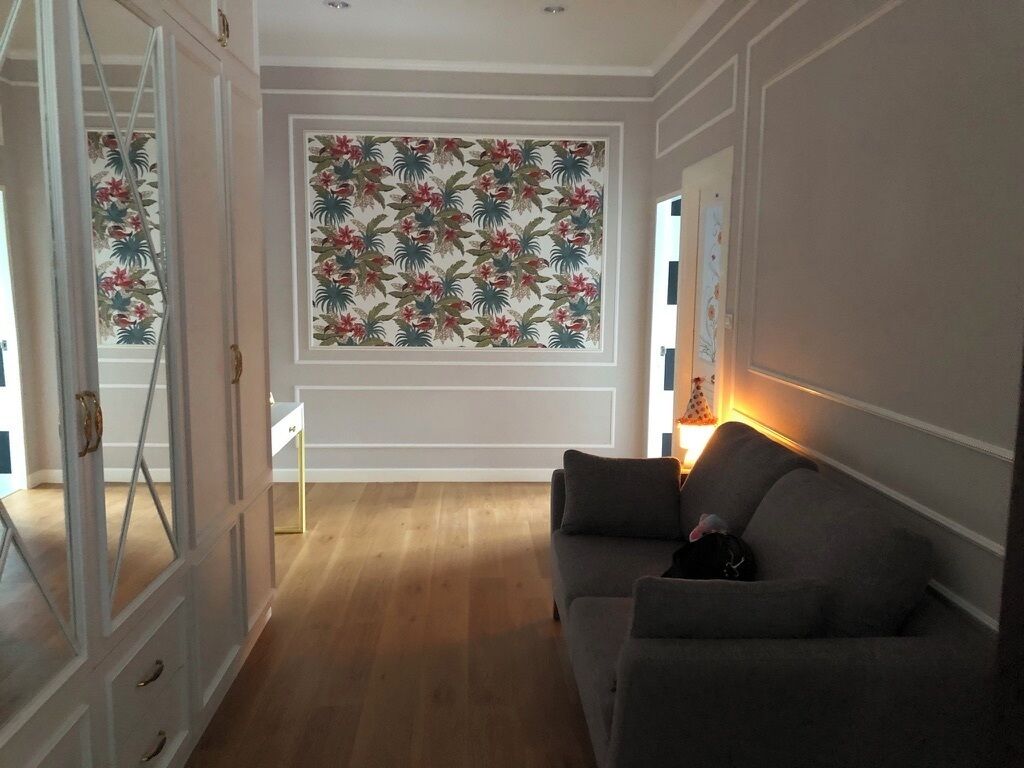 Una habitación con suelo de madera, un sofá gris, una pared con papel tapiz floral y una puerta con marco blanco.