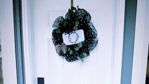 Une porte blanche, ornée d’une guirlande au-dessus. On y trouve un paillasson sur lequel est écrit le mot « MAISON » et un symbole en forme de cœur.