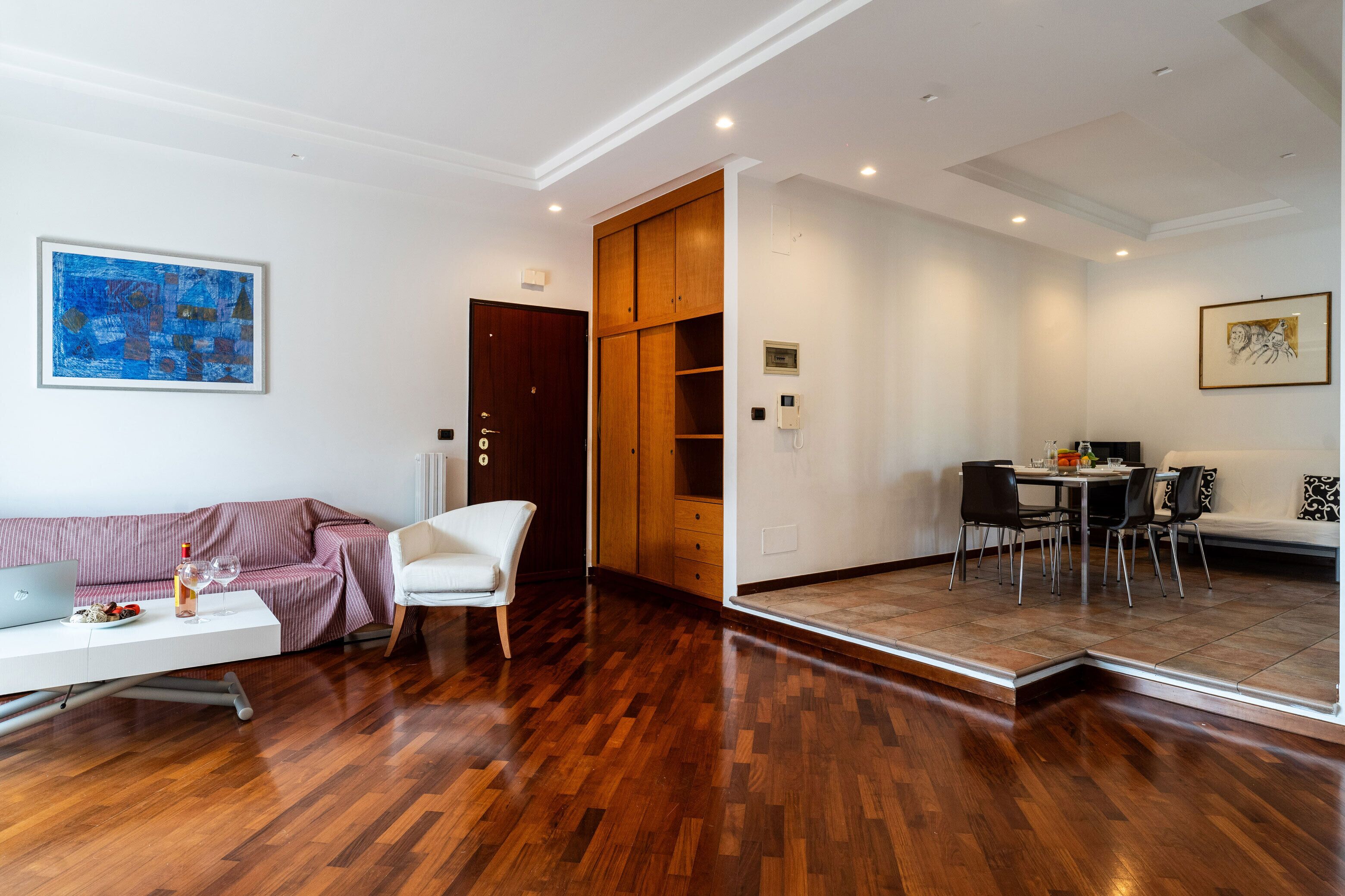 A modern living room with a wooden floor, a white sofa, a white chair, a dining table with chairs, and a wooden cabinet.