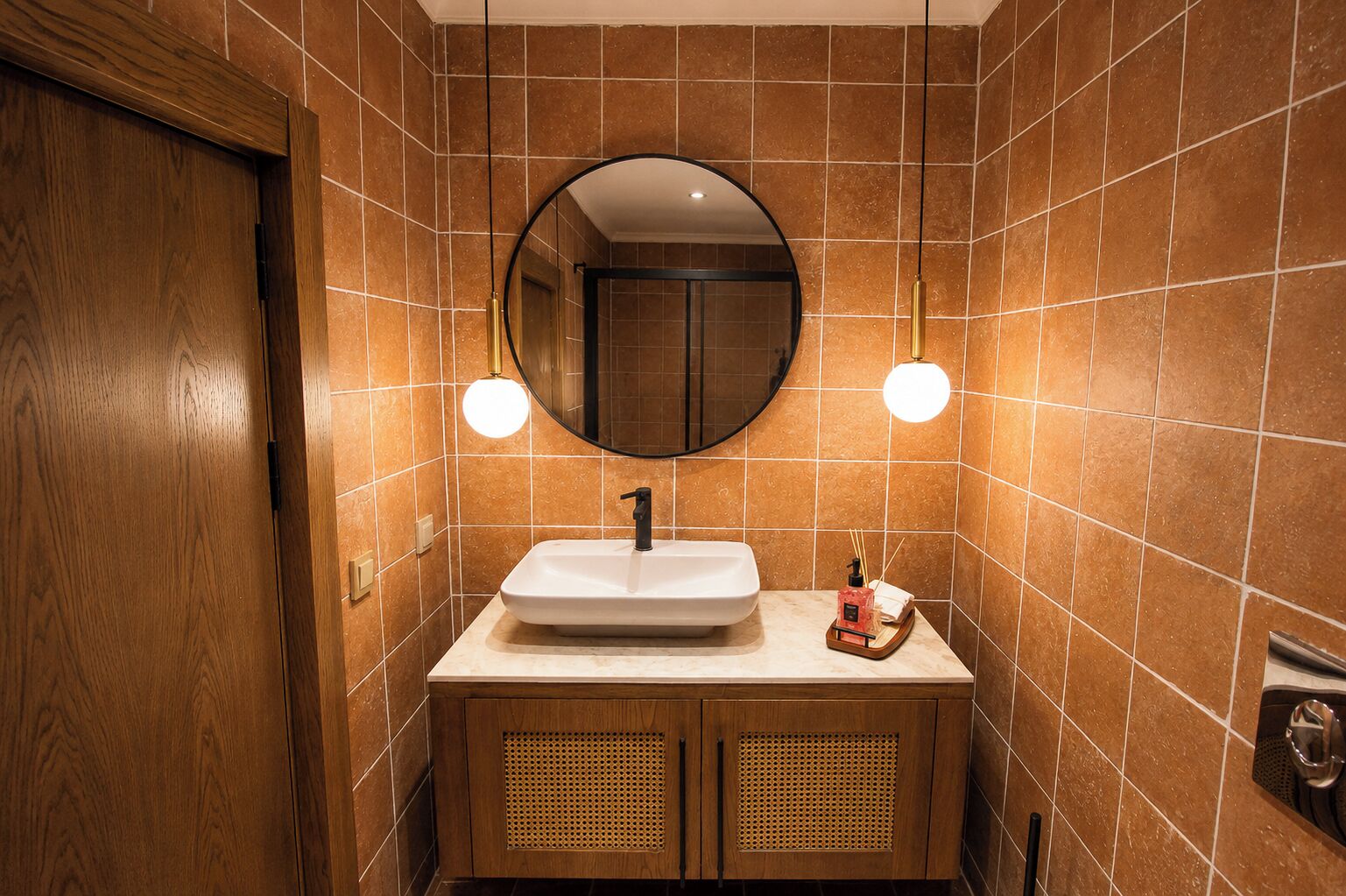 A bathroom with a wooden door, a white sink, a round mirror, and two wall-mounted lights.