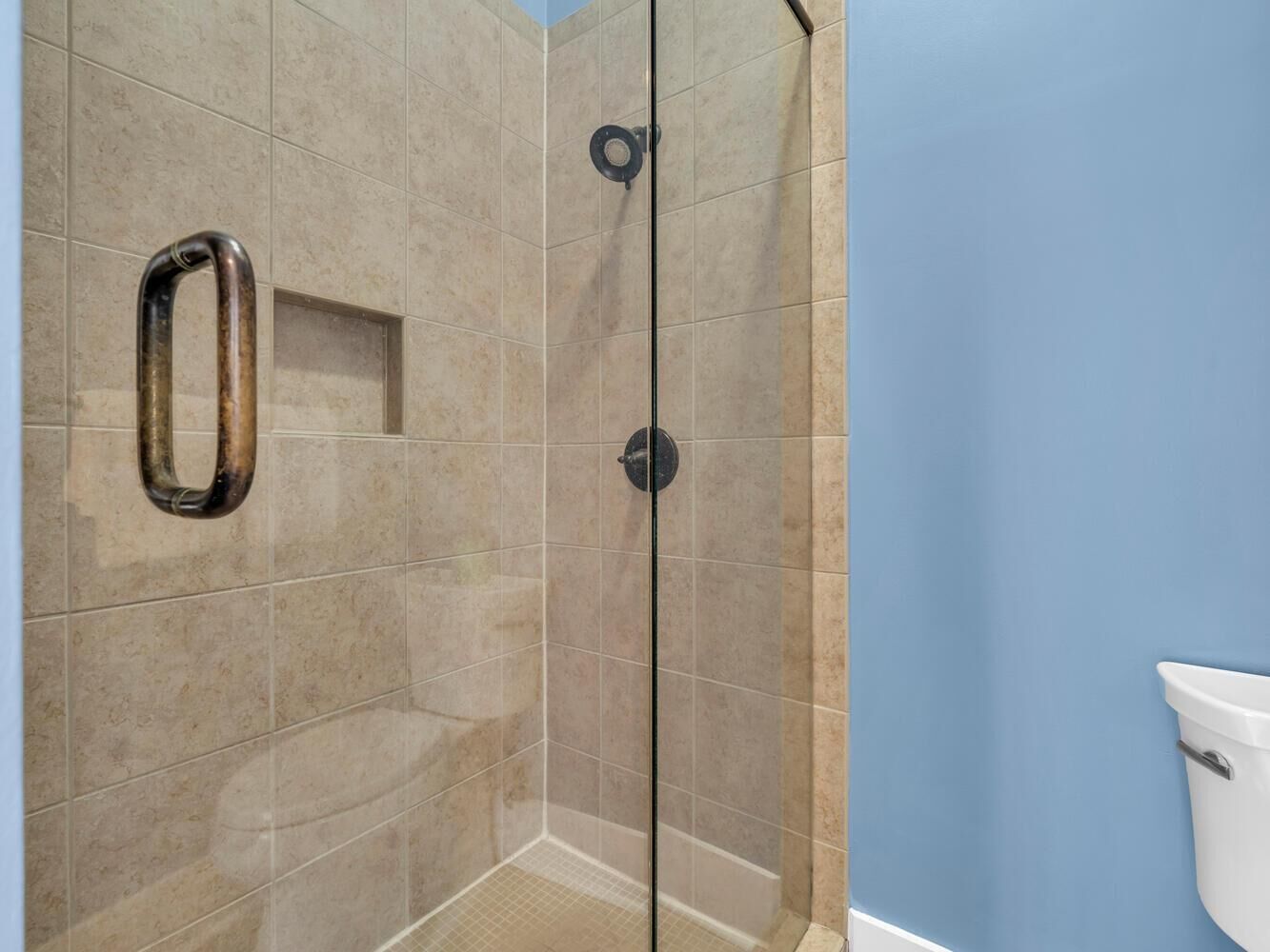Ducha con puerta de vidrio, azulejos color beige y una barra de apoyo metálica.