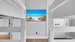 A room with bunk beds, a wall-mounted TV displaying a beach scene, and a wooden floor. - Beach, Please (Santa Rosa Beach)