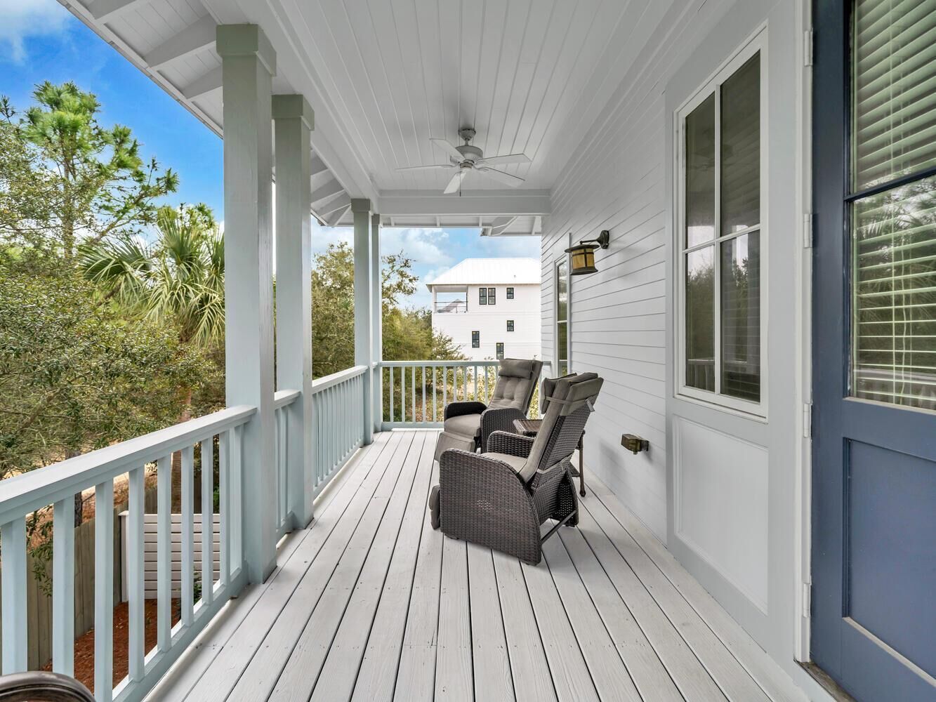A covered porch with two wicker chairs, a ceiling fan, and a view of a white house in the background.