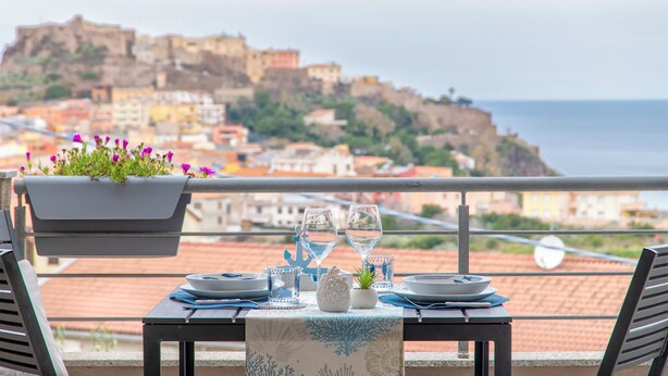 Tavolo apparecchiato per due con vista su una cittadina costiera e un castello sullo sfondo.