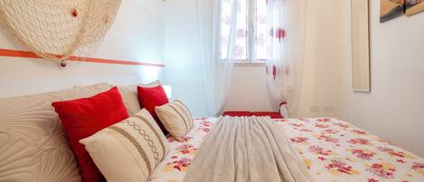 A neatly made bed with floral bedding, a hanging net, and a window with sheer curtains.