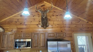 A wooden cabin interior with a mounted deer head, wooden cabinets, a stainless steel refrigerator, and a microwave.
