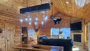 A rustic cabin interior with a wooden dining table, a sofa, a mounted deer head, and a ceiling fan.