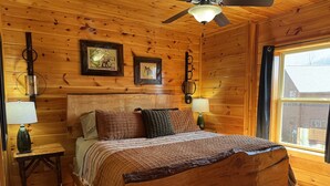 A wooden cabin bedroom with a bed, two wall-mounted lamps, a ceiling fan, and framed pictures on the wall.