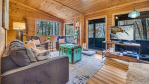 Ein gemütliches Blockhaus-Interieur mit Holzbalkendecke, Deckenventilator, Fernseher, einer Couch und einem großen Fenster.