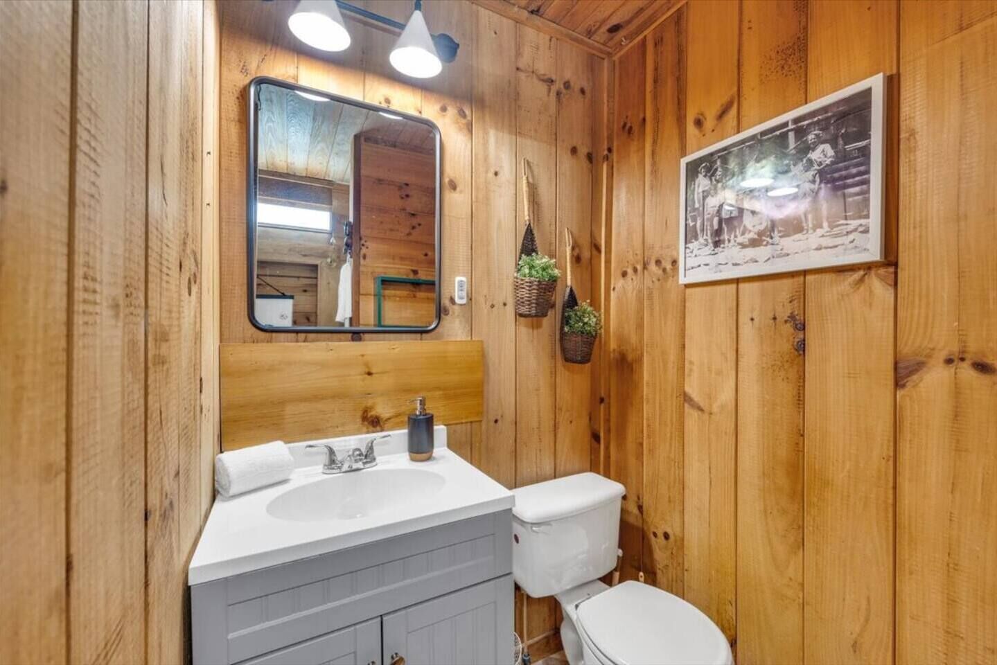 Een badkamer met houten lambrisering, een witte wastafel, een toilet, een spiegel en een ingelijst foto aan de muur.