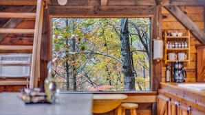 A cabin interior with a large window overlooking a forest, a kitchen area with wooden cabinets, and a bar counter.