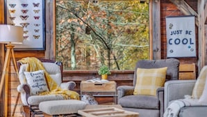 A cozy cabin interior with a large window, a framed butterfly collection, and a 'Just be cool' sign.