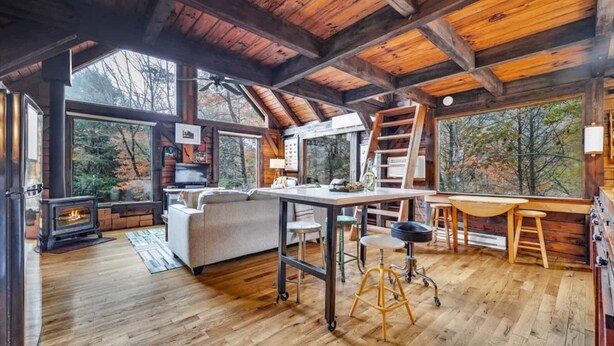 A cozy cabin interior with a wooden ceiling, a fireplace, a sofa, a dining table, and large windows offering a view of trees.