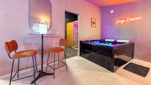 Une pièce avec une table de baby-foot, deux tabourets de bar, un miroir et un panneau néon sur lequel on peut lire : « Deep Desire ».