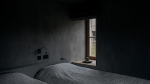 A dimly lit room with a single bed, a small table, and a window with a view of the outdoors.