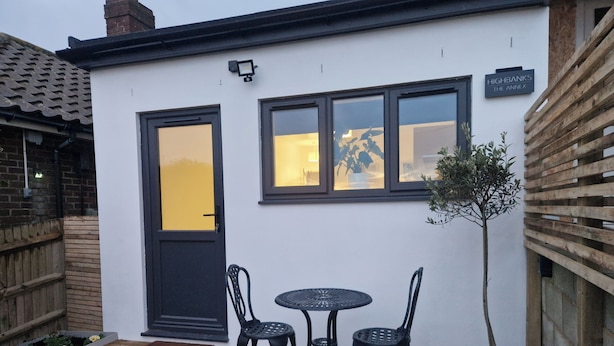 A white building with a black door and windows, a small patio with a table and chairs, and a potted plant. - Cozy Apartment at Highbanks The Annex (Newhaven)