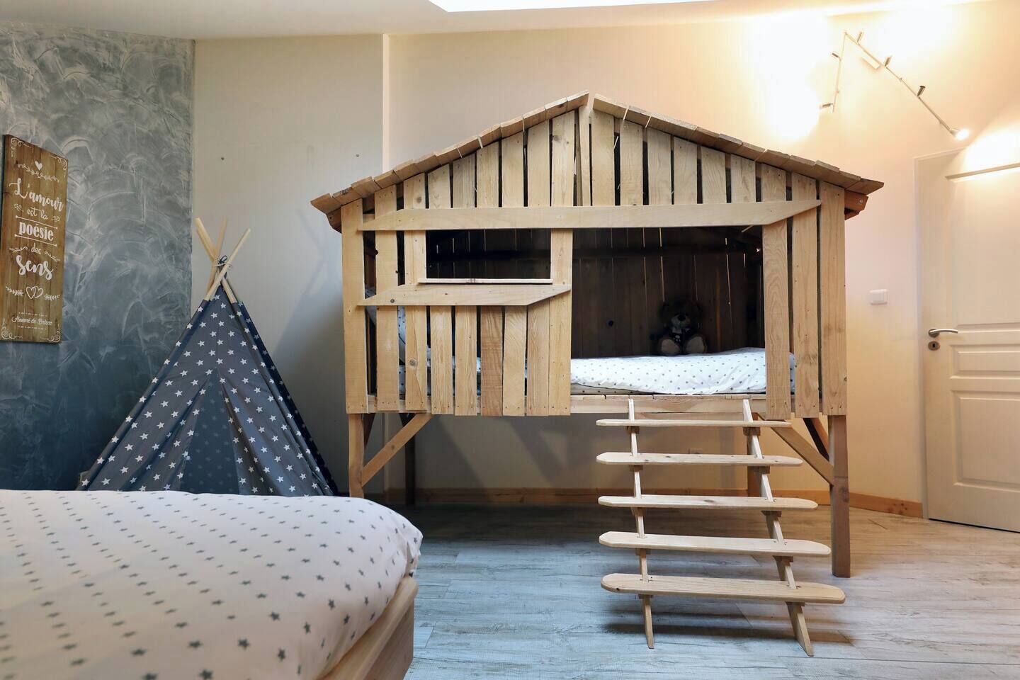 Une chambre à coucher accueillante avec un lit superposé en bois, une petite fenêtre, une échelle, une tente et un mur orné d’une enseigne.