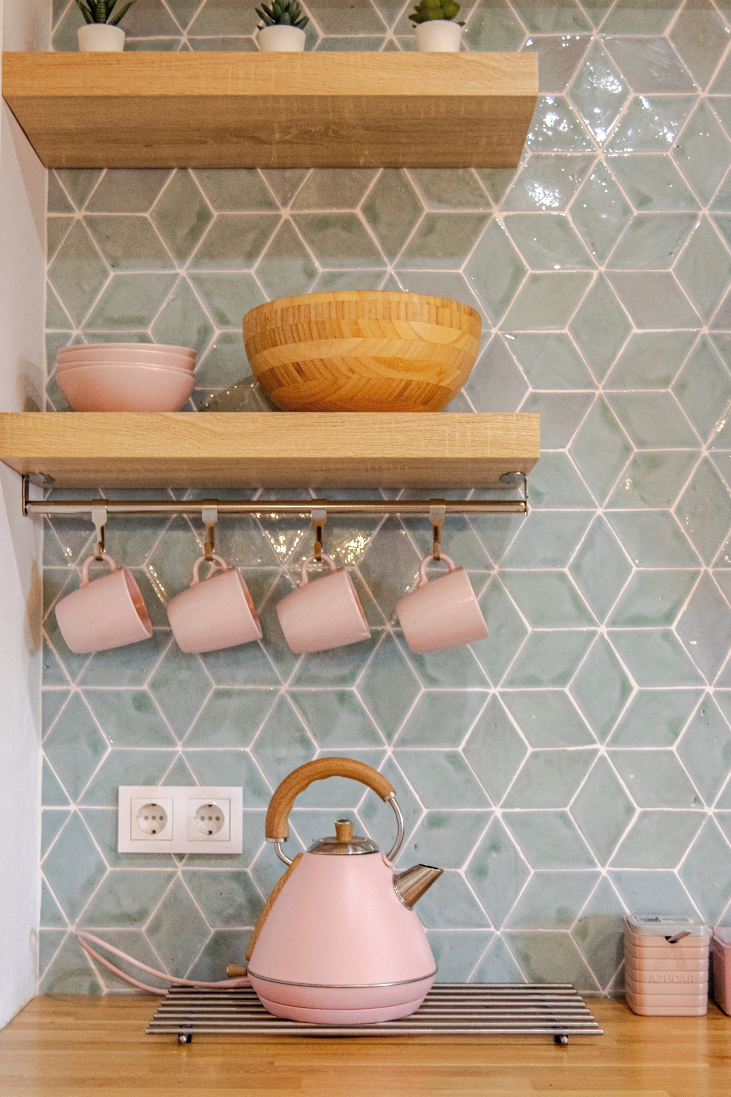 A kitchen with a wooden shelf, hanging pink cups, a pink kettle, and a hexagonal tiled backsplash.