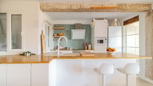 Een moderne keuken met een houten aanrecht, witte kasten en een centraal eiland met barkrukken. Er hangen hanglampen, een magnetron en een schaal met fruit op het eiland.
