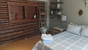 A log cabin-style bedroom with a wooden ladder, a bedside table with a lamp, a stack of towels, and a bookshelf with books and decorative items.