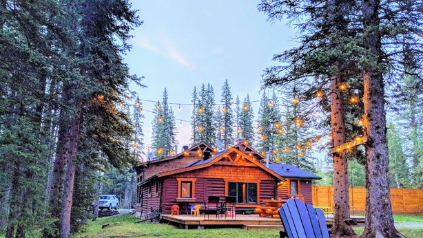 A log cabin with a deck and string lights, surrounded by tall trees.