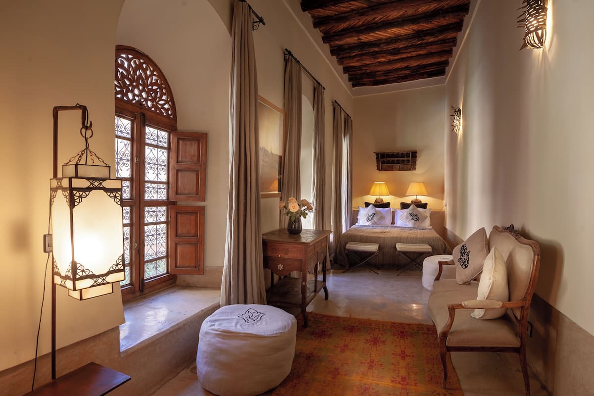 A cozy room with a wooden ceiling, a sofa, a chair, a small table with a vase, and a bed with two lamps.