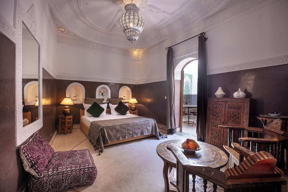 A traditional room with a bed, a sitting area with a table and chairs, a chandelier, and a wooden cabinet.