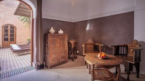 A traditional Moroccan room with a patterned floor, a wooden cabinet, a round table with fruit, and a view of an outdoor area with a brick wall and a wooden door. - RIAD BOHEMIA (Marrakech)