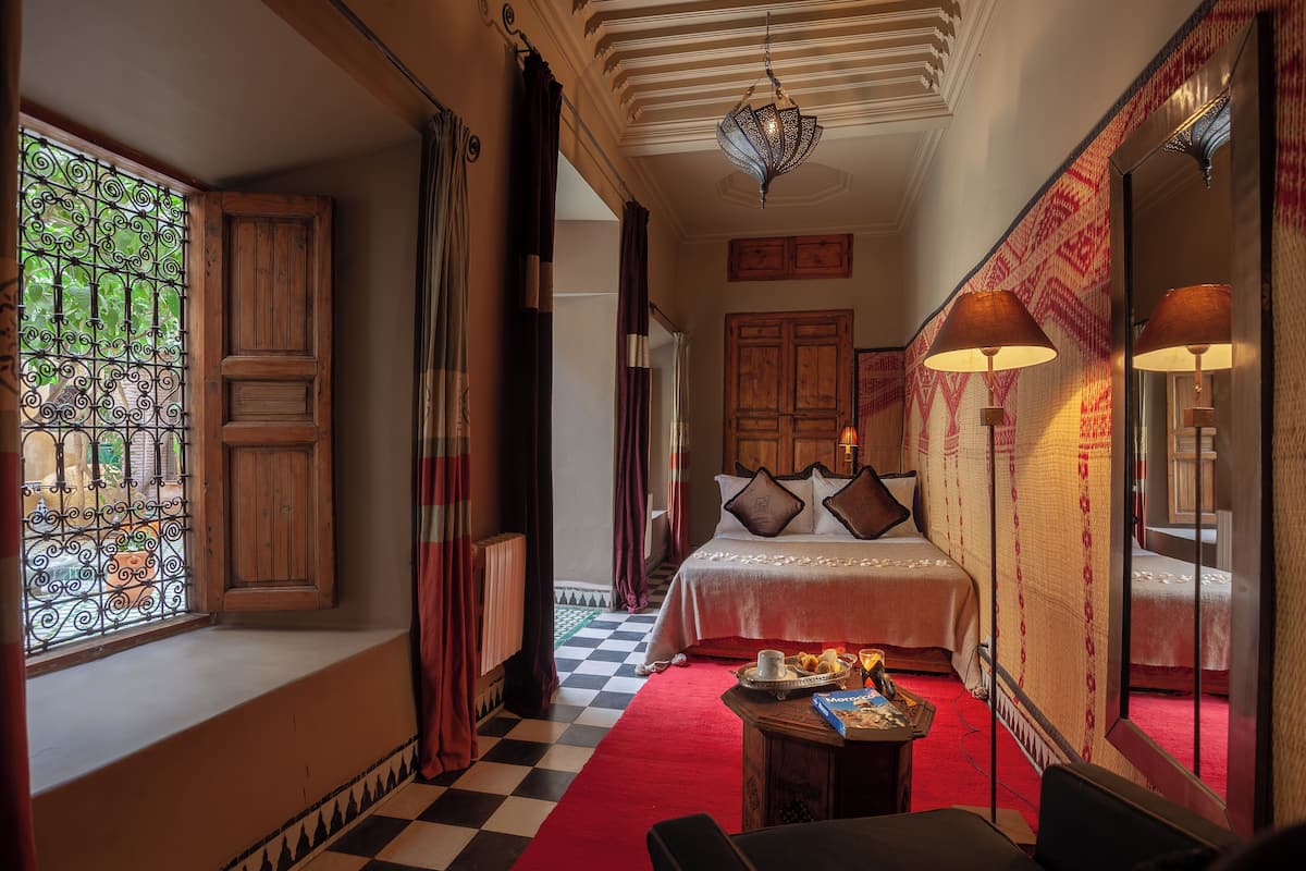A hotel room with a checkered floor, a bed with pillows, a table with a tray, and a window with curtains.