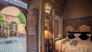 A bedroom with a bed, a nightstand, a lamp, and a view of an outdoor courtyard with a fountain and plants. - RIAD BOHEMIA (Marrakech)