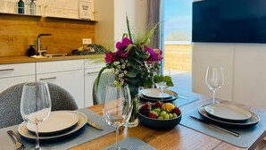 A dining area with a table set for two, a fruit bowl, a vase of flowers, and a view of the outdoors.