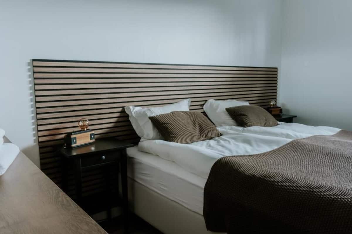 A neatly made bed with white linens and two brown pillows, a wooden headboard, a bedside table with a clock, and a small nightstand with a lamp.