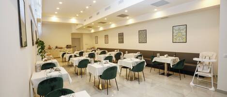 A spacious dining area with multiple tables and chairs, a high ceiling with recessed lighting, and framed artwork on the walls.