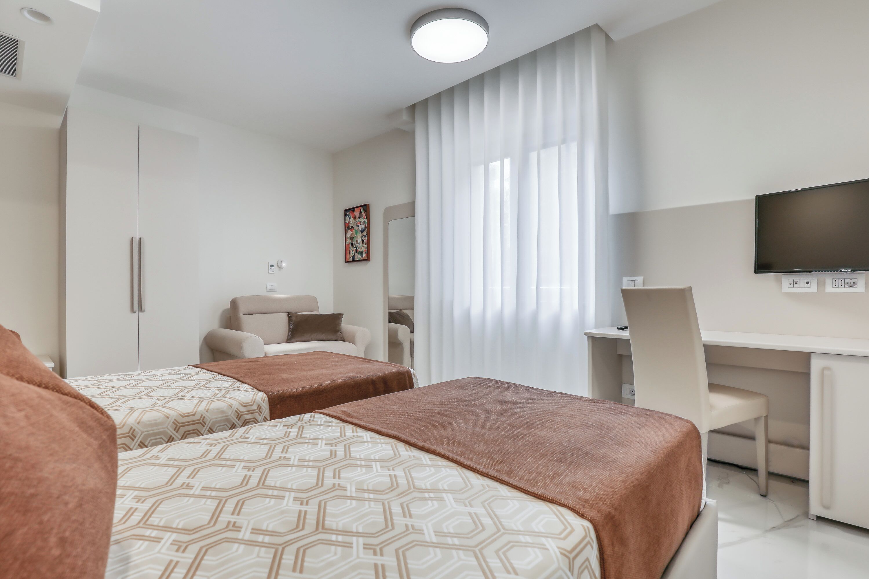 A hotel room with two beds, a desk, a chair, a TV, and a window with curtains.