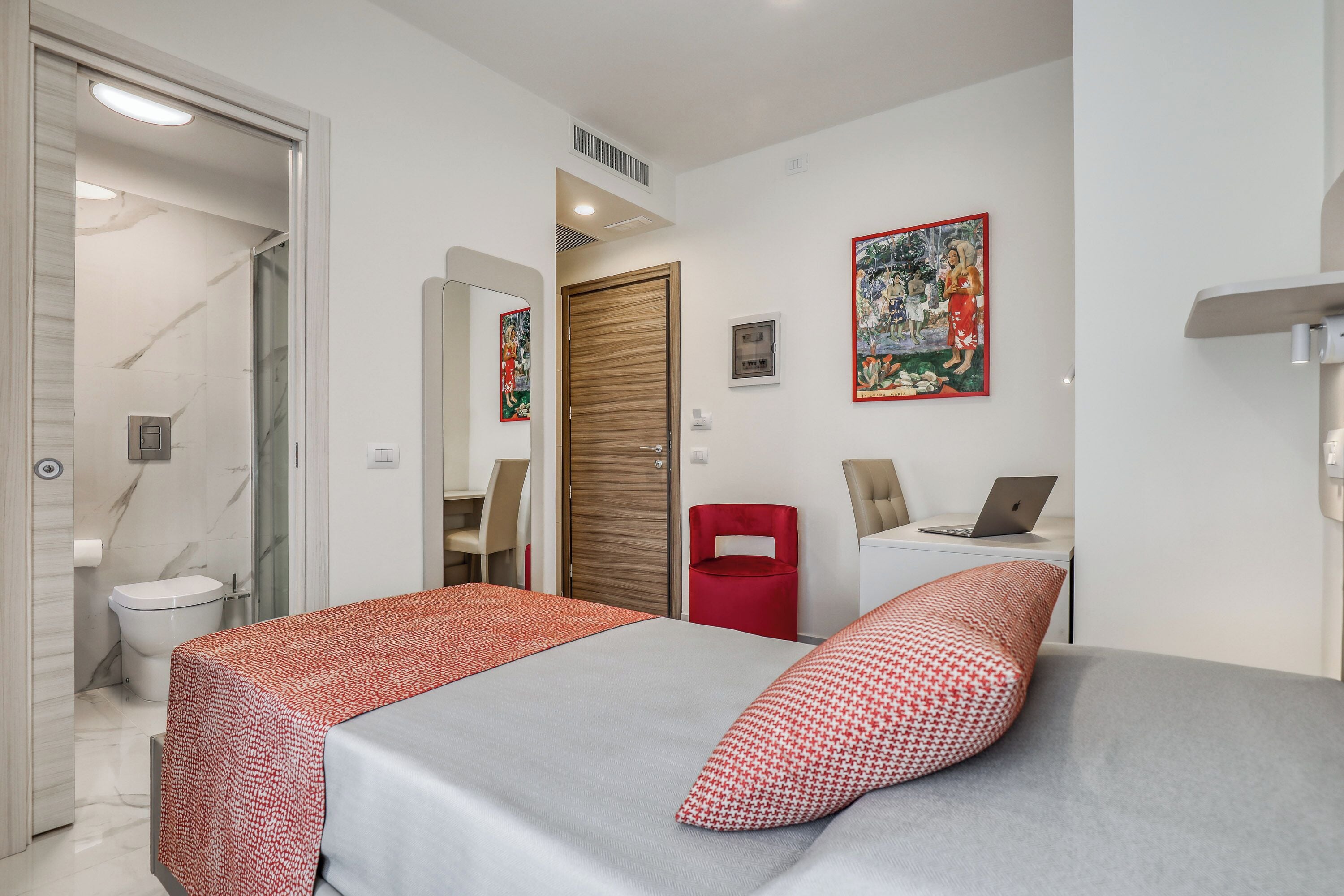 Une chambre d’hôtel comprenant un lit, un bureau avec un ordinateur portable, une chaise rouge, des toilettes et un espace douche.