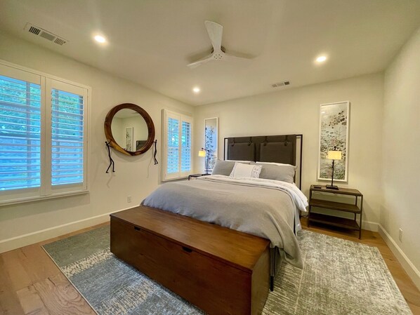 A bedroom with a bed, a bedside table, a nightstand, a floor lamp, a ceiling fan, and a large window with blinds.