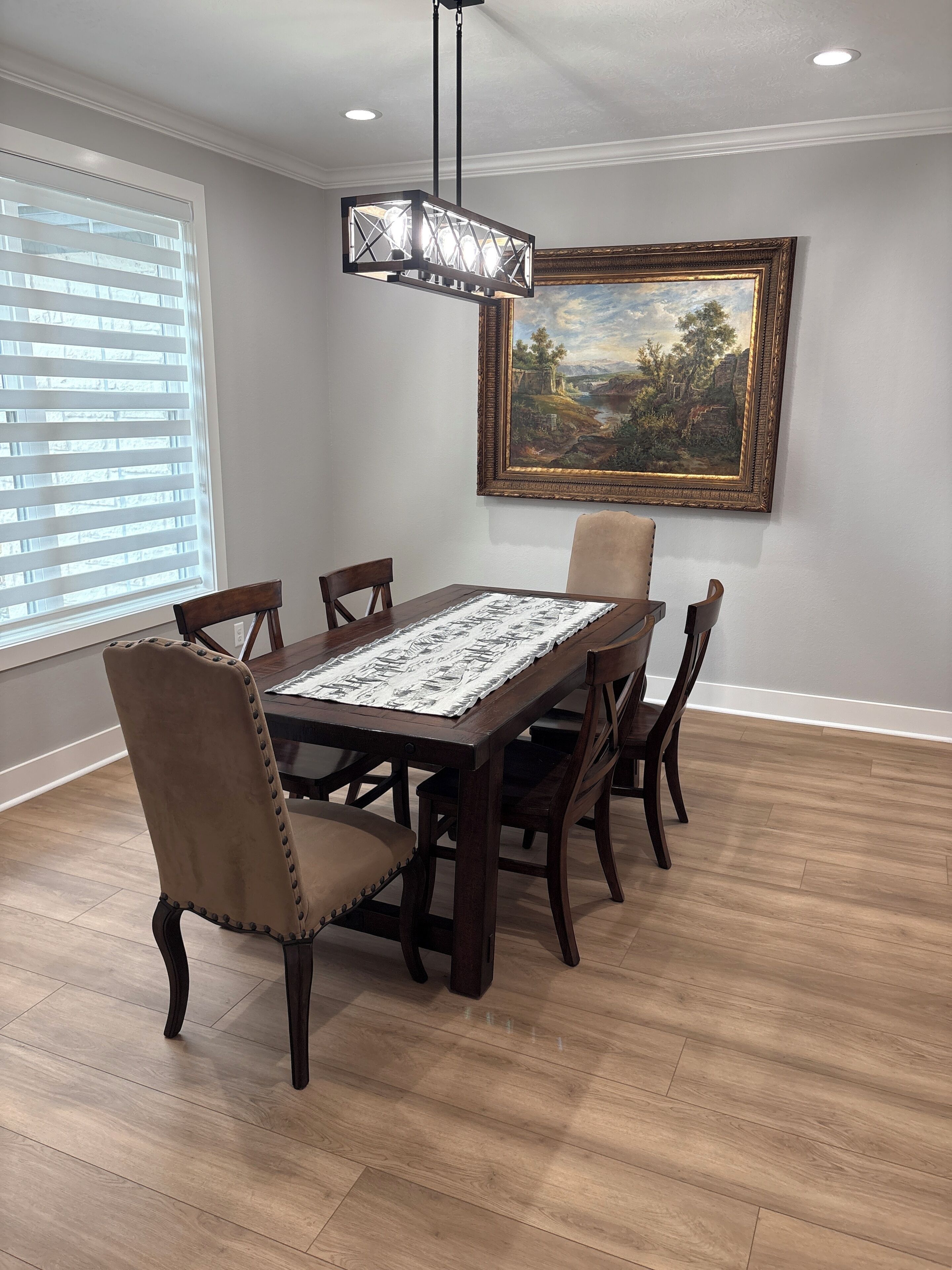A dining room with a wooden table, six chairs, a chandelier, a painting on the wall, and a window with blinds.