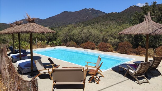 Uma área de piscina com espreguiçadeiras e toldos de palha, rodeada por vegetação exuberante e montanhas.