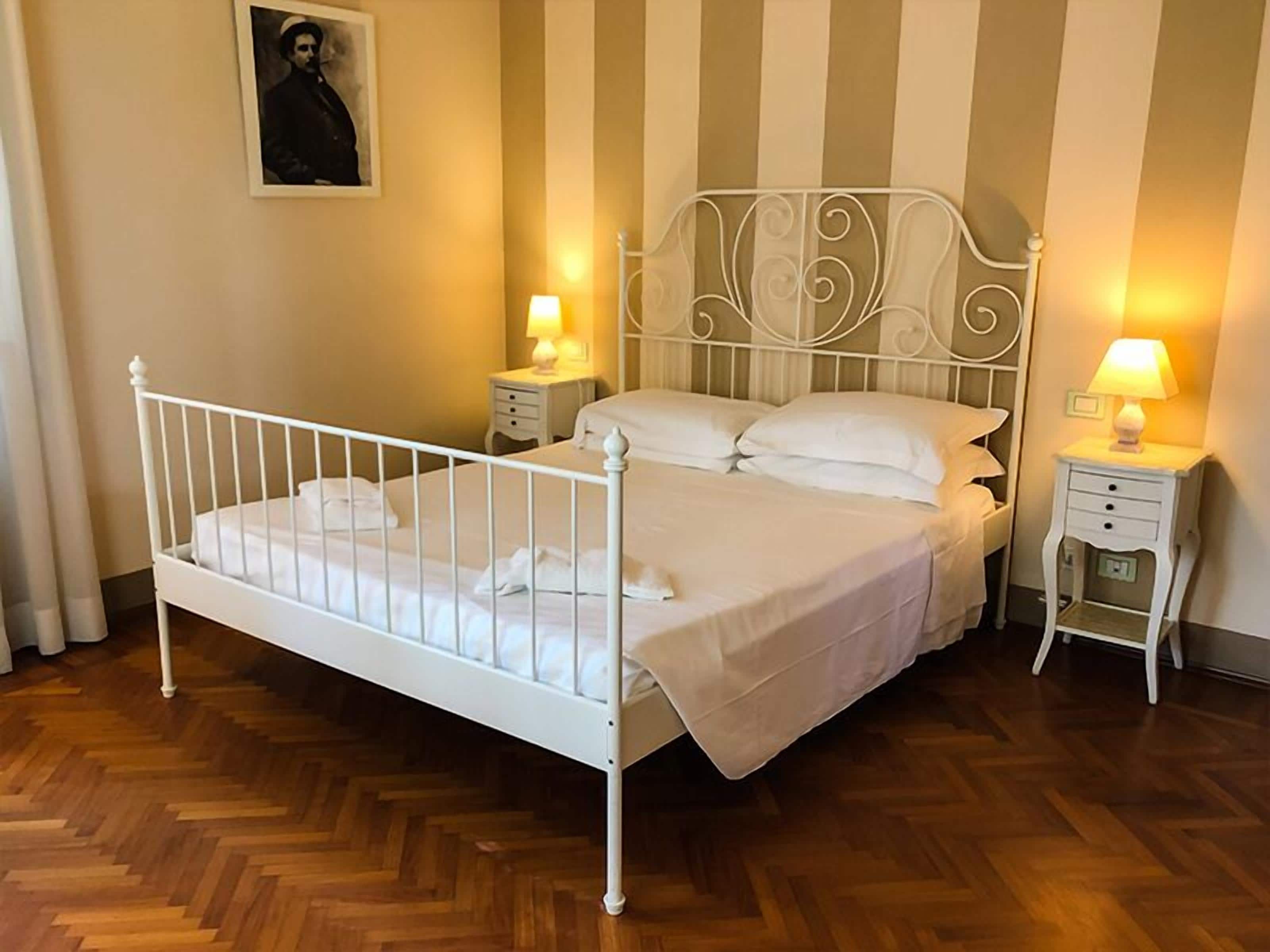 Una cama bien hecha con ropa blanca, dos mesitas de noche con lámparas y una pared a rayas al fondo.