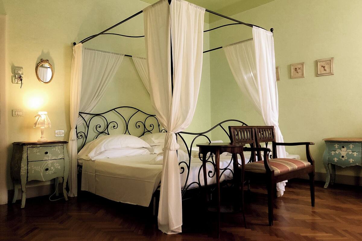 A four-poster bed with white curtains, a wooden chair, a small table, and a nightstand with a lamp.