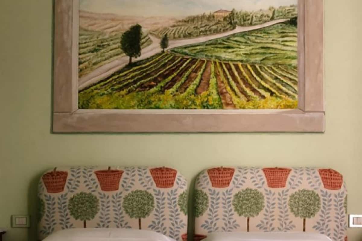 A room with a bed, two pillows with a basket and tree pattern, and a painting of a field on the wall.