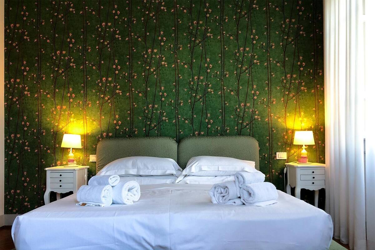 A neatly made bed with white linens and towels, two bedside tables with lamps, and a green patterned wall.