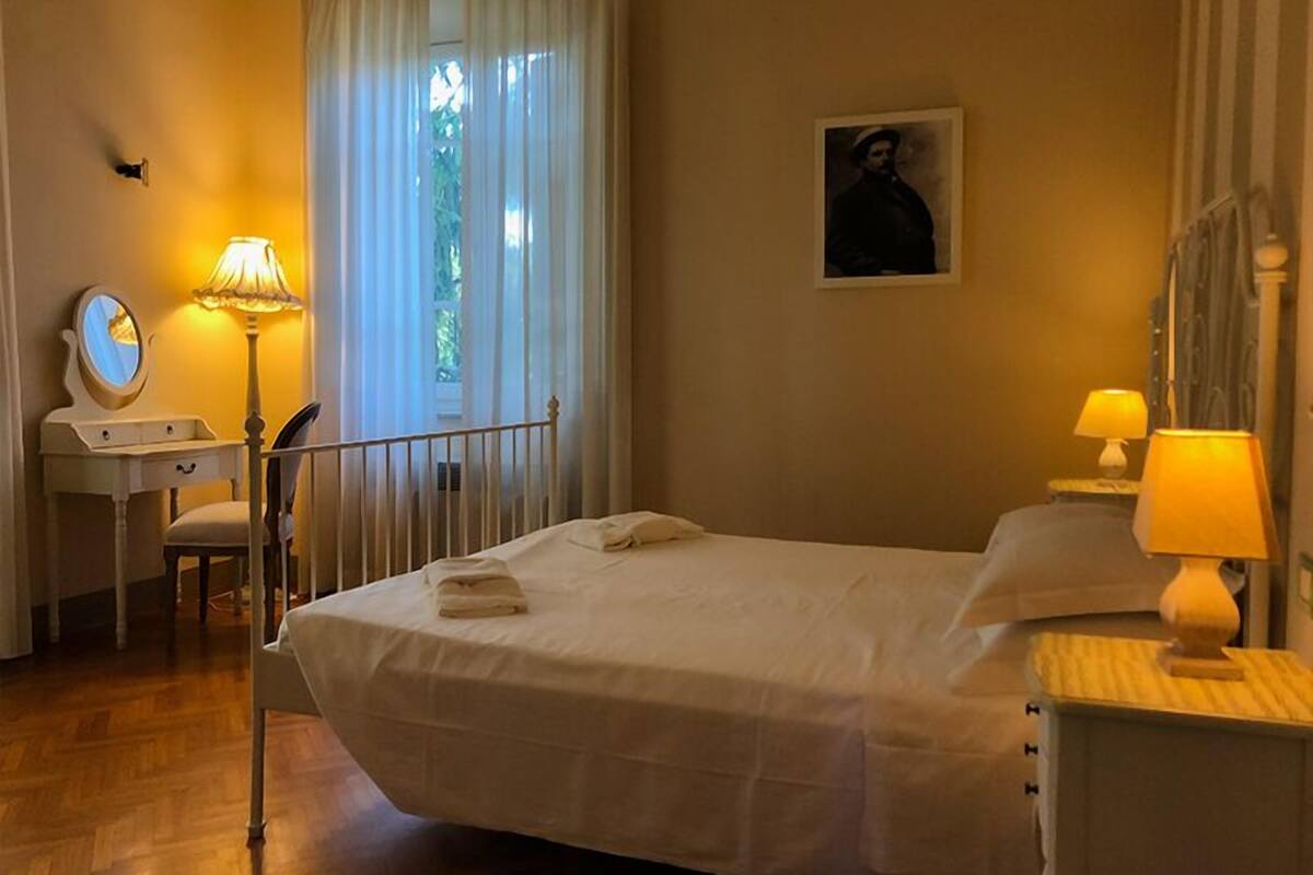 A neatly arranged bedroom with a bed, bedside lamps, a desk with a mirror, and a window with curtains.