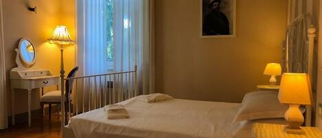 A neatly arranged bedroom with a bed, bedside lamps, a desk with a mirror, and a window with curtains.