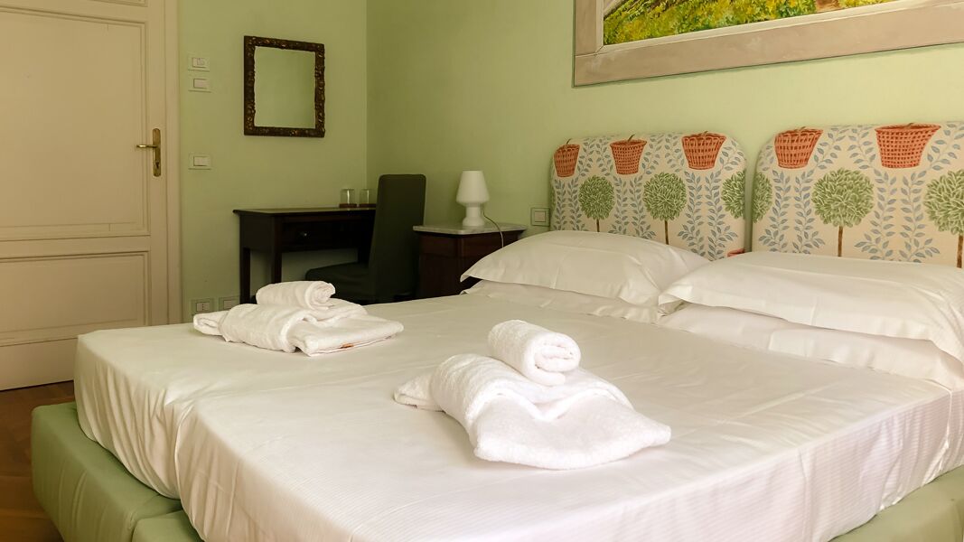 A hotel room with a bed, two rolled towels, a desk, a chair, a lamp, and a wall with a mirror and patterned headboard.